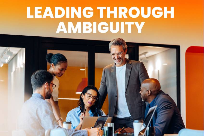 Diverse team collaborating around a table with laptops under the headline “Leading Through Ambiguity.”