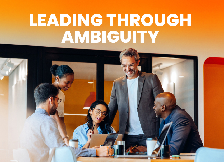 Diverse team collaborating around a table with laptops under the headline “Leading Through Ambiguity.”